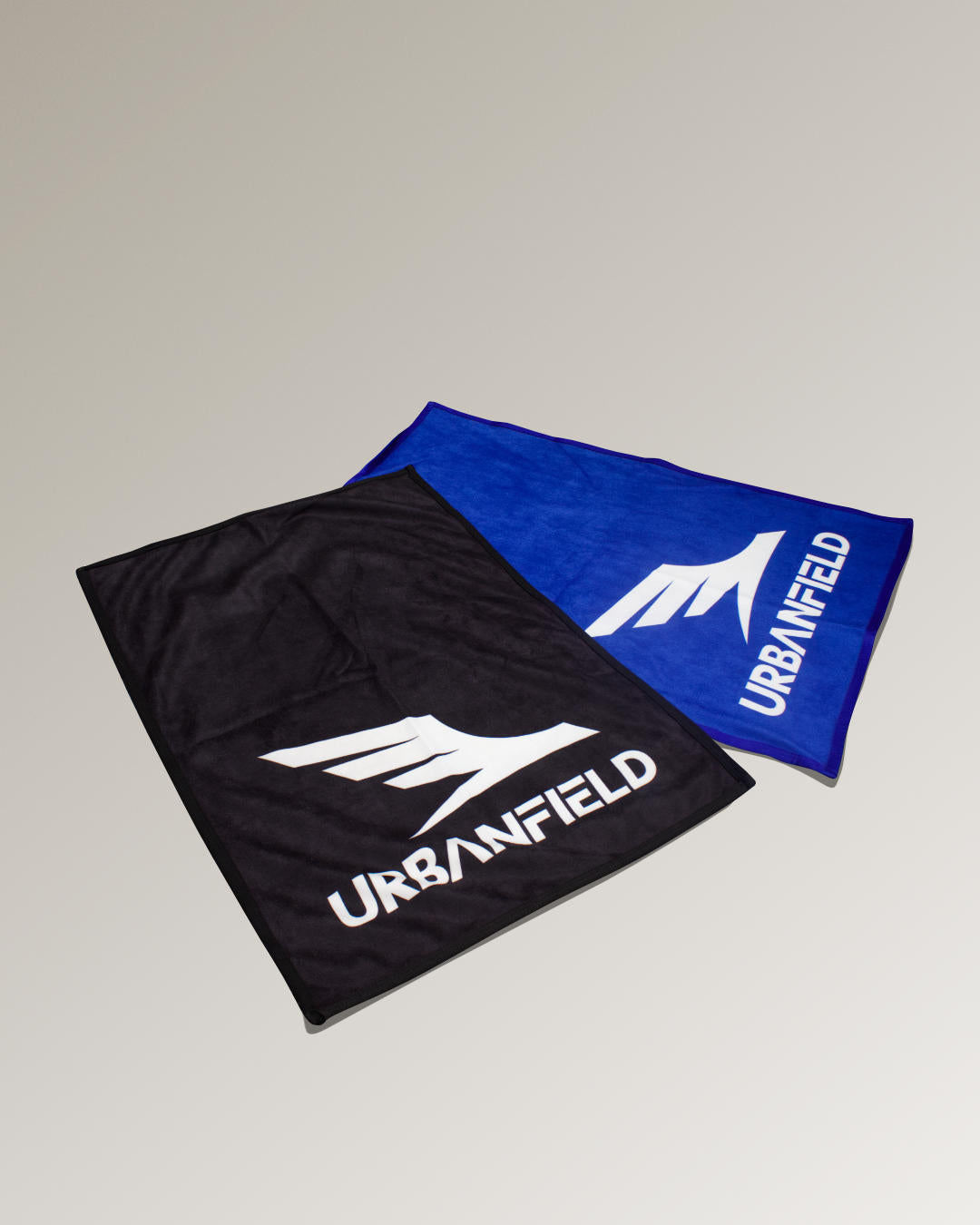 Two black and blue Urbanfield Sports logo hand towels on a dark beige background.