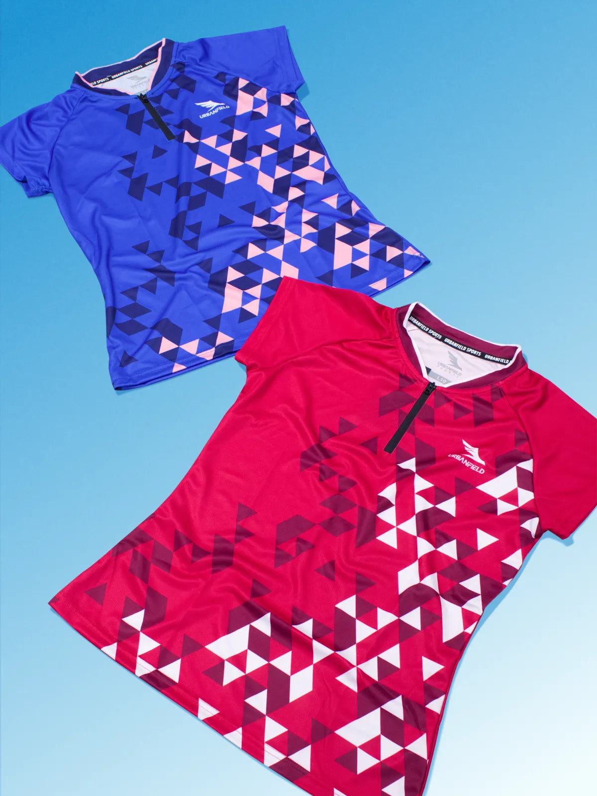 Two women's Urbanfield Sports Challenger t-shirts, one in cherry red and the other in pacific blue on a light blue background.