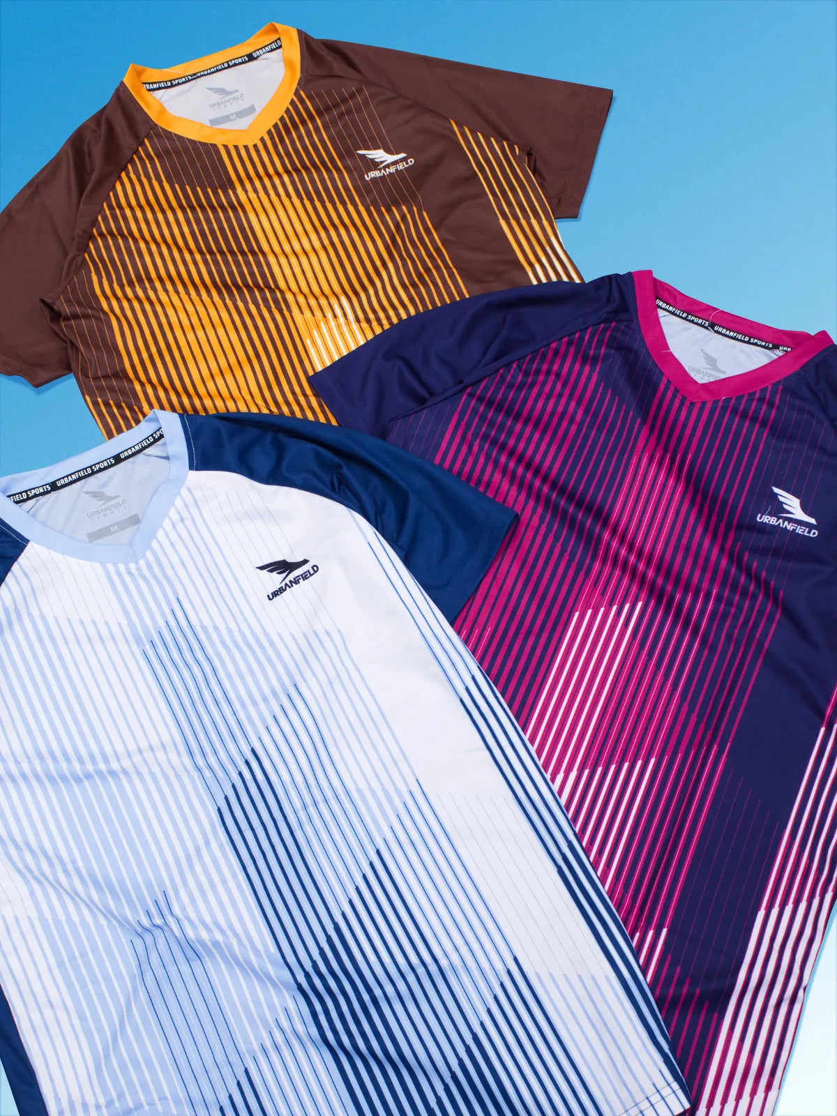 Three men's Urbanfield Sports Ignis t-shirts, in slate blue, navy and brown on a light blue background.