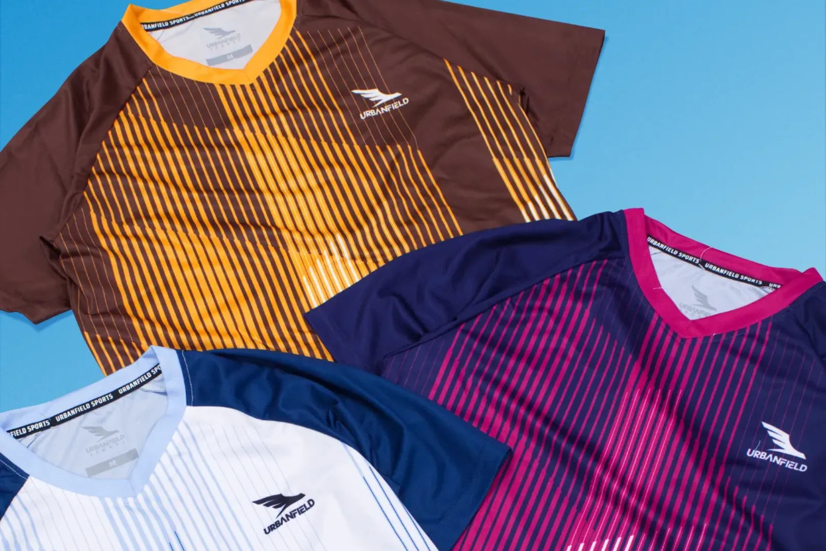 Three men's Urbanfield Sports Ignis t-shirts, in slate blue, navy and brown on a light blue background.
