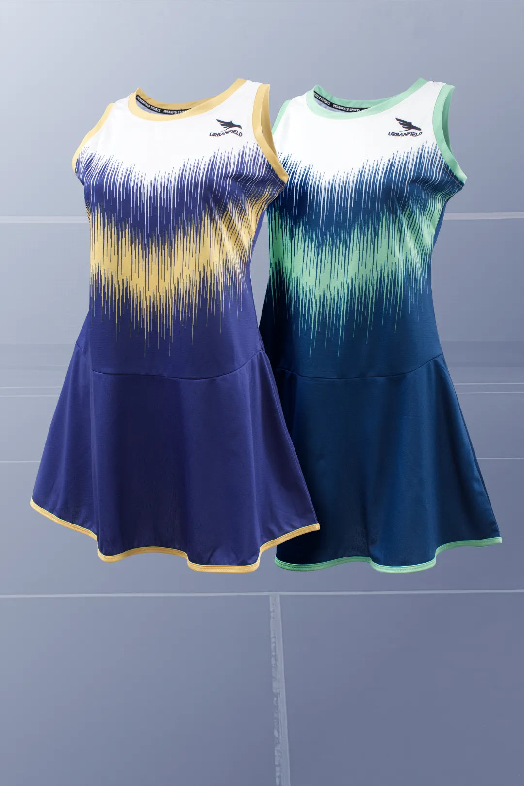 Two Urbanfield Sports women's striker sleeveless dresses, one in carbon navy and the other in slate blue, are displayed against a grey badminton court background.