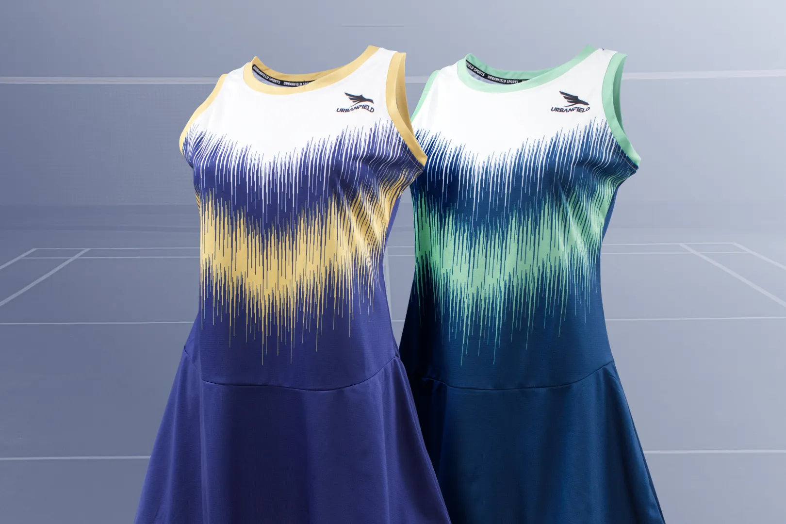 Two Urbanfield Sports women's striker sleeveless dresses, one in carbon navy and the other in slate blue, are displayed against a grey badminton court background.